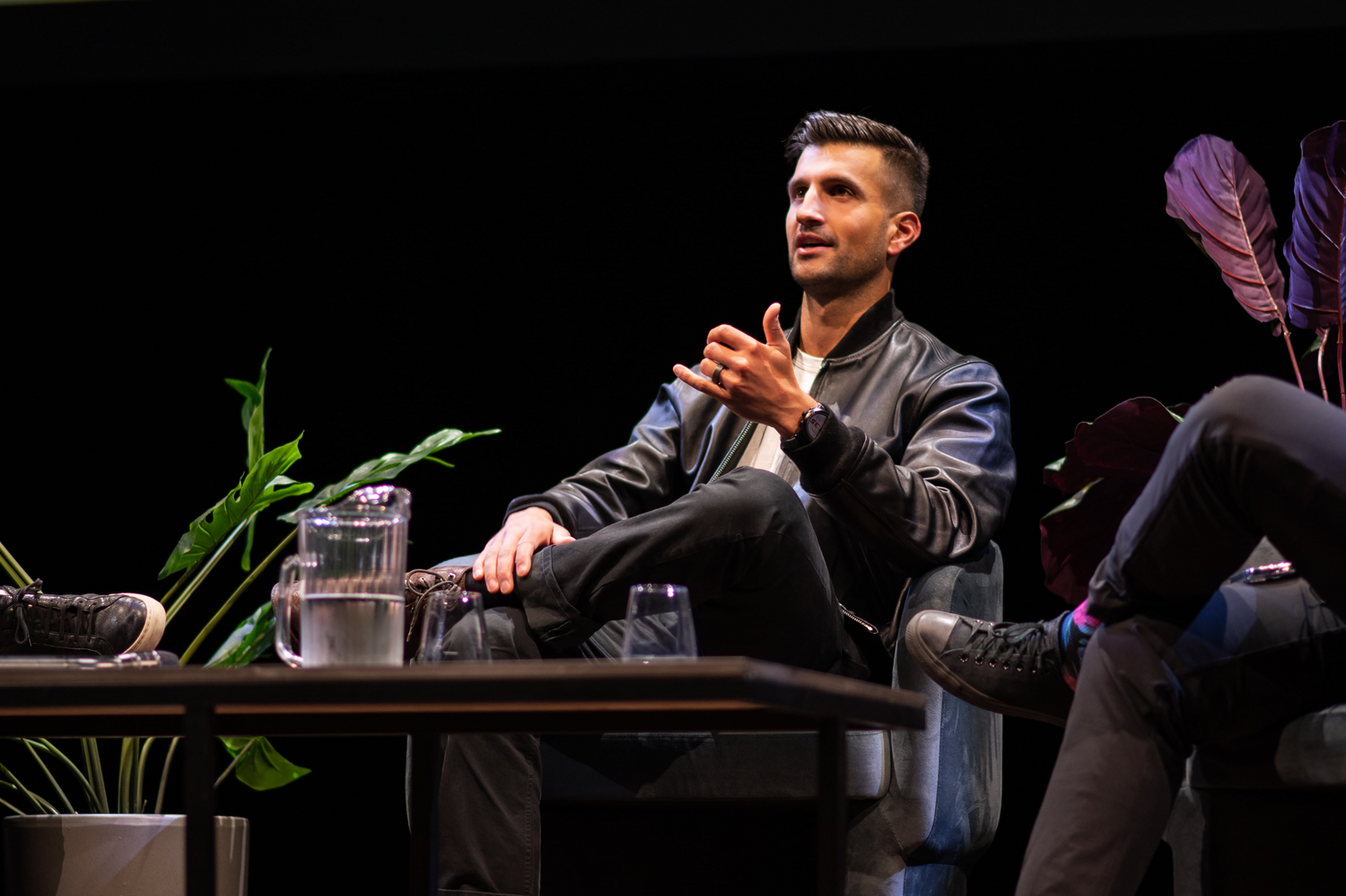 Man sitting on a stage with a dark background, speaking or listening.