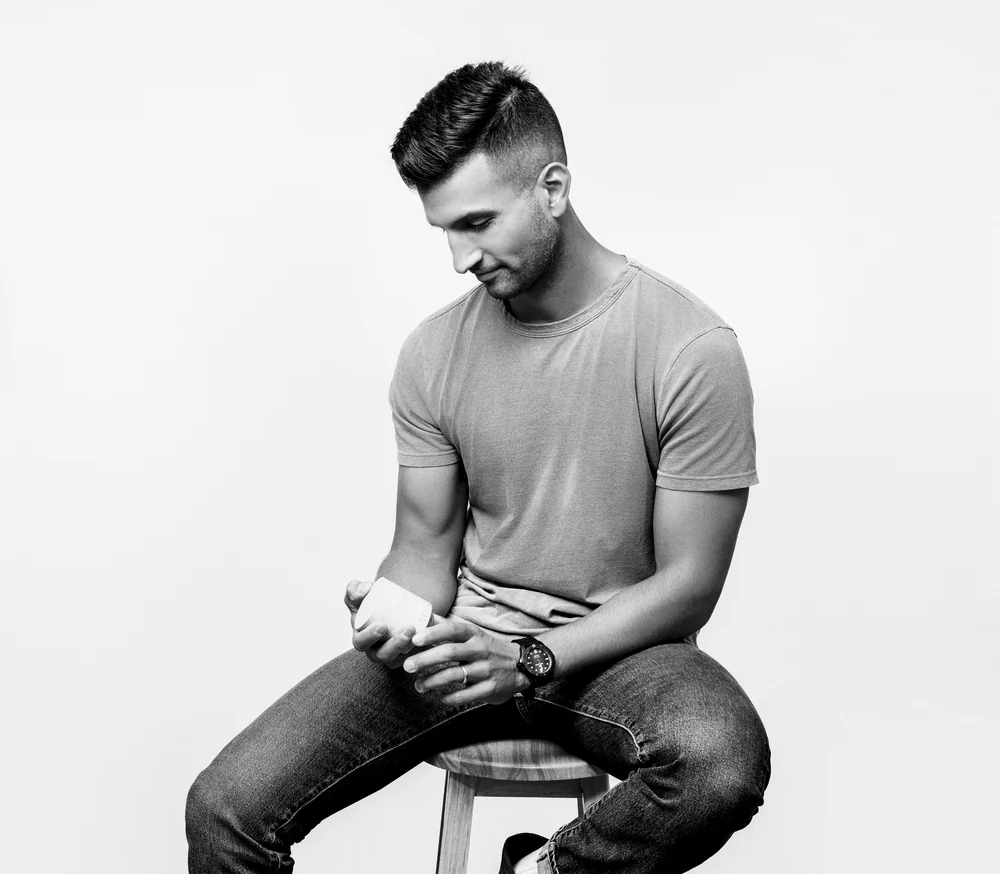 Wild Roman founder Sahil Bloom wearing a short-sleeve crewneck t-shirt and jeans sits on a stool against a plain light background, captured in a black and white photograph.
