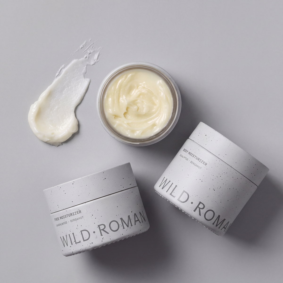 Overhead view of three Wild Roman moisturizer jars, one open showing the cream, with a cream swatch