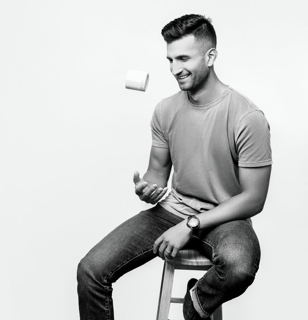 A man sitting on a stool in a studio setting, wearing a light gray t-shirt and dark jeans, with a white cylindrical product floating near his head.