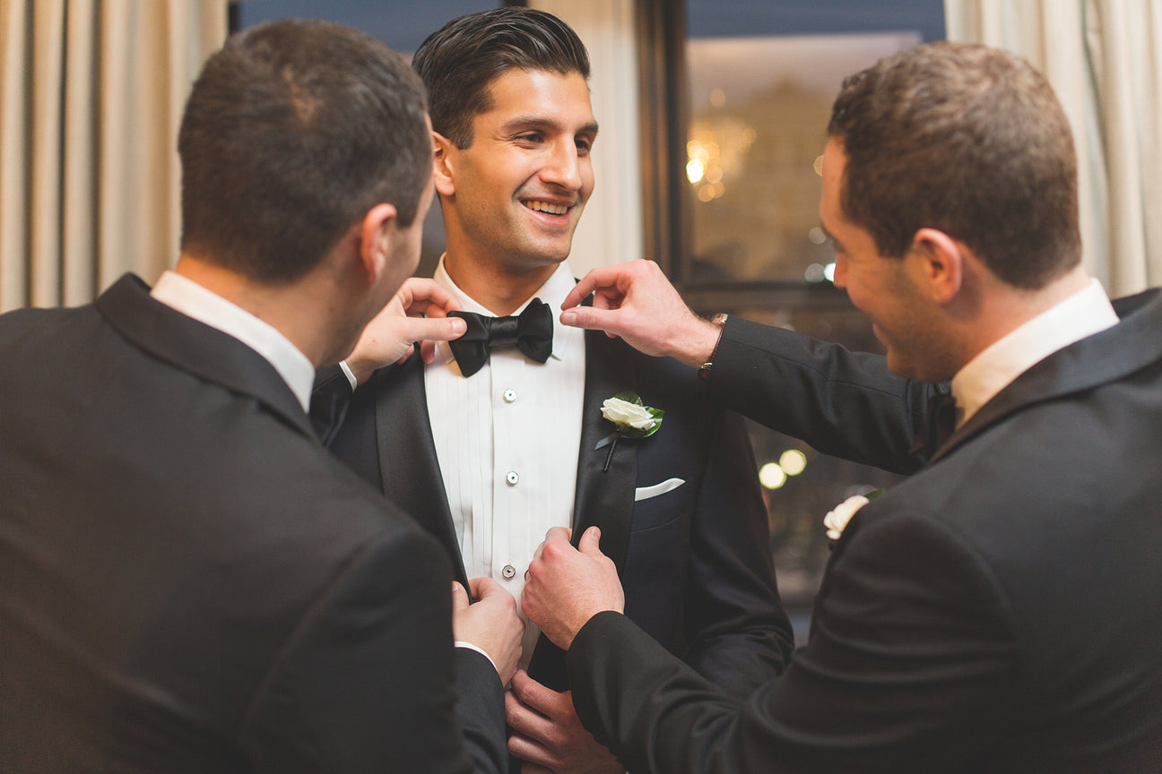 A smiling groom in a black tuxedo jacket with a white shirt and black bow tie, having his suit adjusted by two groomsmen in black tuxedos.