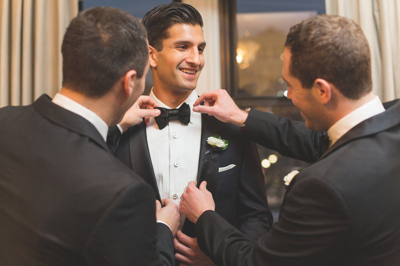 A smiling groom in a black tuxedo jacket with a white shirt and black bow tie, having his suit adjusted by two groomsmen in black tuxedos.